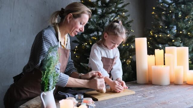 A happy mother, a young woman, cooks Christmas cookies with her little daughter, a cute girl sitting at a table in a house kitchen