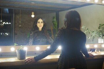 A beautiful young woman stands near a mirror with light bulbs in a room with a loft interior