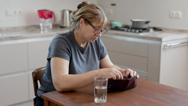 A Caucasian woman is sitting at kitchen and cannot find money in her wallet. Poor woman with empty wallet sitting at home.