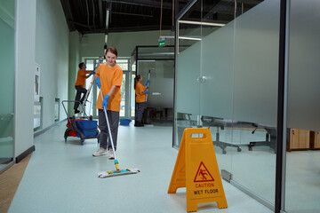 Young woman wiping floor with wet mop near warning sign