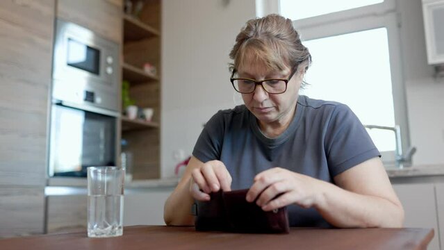 A Caucasian woman is sitting at kitchen and cannot find money in her wallet. Poor woman with empty wallet sitting at home.