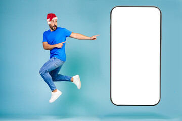 Fototapeta na wymiar Full length portrait of man jumping in blue t-shirt and christmas hat pointing with finger at big screen with poor screen on blue background