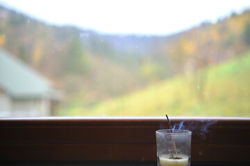 An incense stick smokes while standing in a glass on the windowsill. Outside the window are mountains with autumn doors and a beautiful landscape