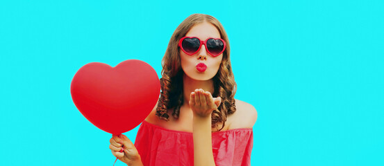 Portrait of beautiful young woman with pink heart shaped balloon blowing her lips sending sweet air kiss isolated blue background