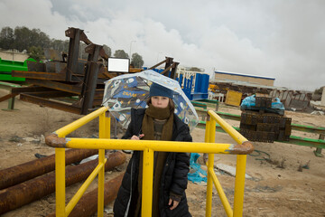 cool stylish boy in a coat and hat with a scarf posing plays and walks through pipes with an umbrella in an industrial area among green yellow blue metal structures against a cloudy sky on a rainy day