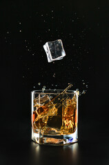 An ice cube falls into a glass of poured whiskey causing a splash and splashes