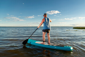 A man in shorts standing on a SUP board with a paddle floats on the water in the rays of the setting sun.