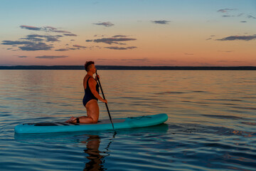 Fototapeta na wymiar a Jewish feminist woman in a closed bathing suit with a mohawk on a koyen on a stand-up board with an oar floats on the water against the background of the sunset sky.