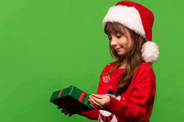 A cute little girl in a Santa hat unpacks a New Year's gift. Merry Christmas and Happy New Year 2023. Portrait isolated on green background