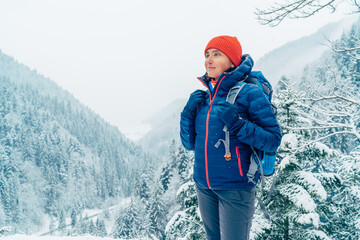 Female backpacker with backpack dressed warm down jacket enjoying snowy mountains landscape while she trekking winter mountain forest route. Active people in the nature concept image.