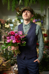 Fototapeta premium Attractive cheerful caucasian male in peaked cap and vintage clothes from 20s holding beautiful bouquet of red and violet flowers. Mother's Day or Valentine's Day concept. High quality image