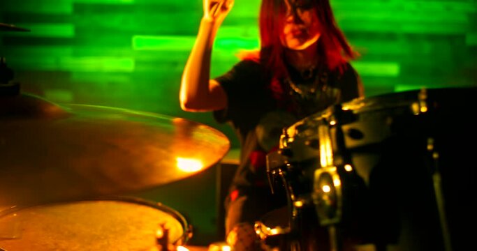 Drummer girl vigorously playing drums in green and orange flickering strobe lights on the nightclub stage. Performance of female rock band. Focus shifts from the drum set to the girl and back again.