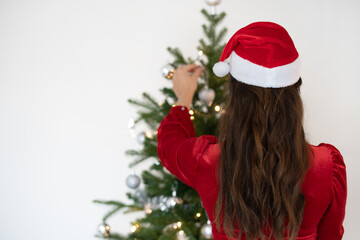 Nahaufnahme von einer Frau von hinten, mit einer roten Weihnachtsmütze auf dem Kopf, die einen Weihnachtsbaum an Weihnachten schmückt. Weihnachtlicher Hintergrund mit Platzhalter.