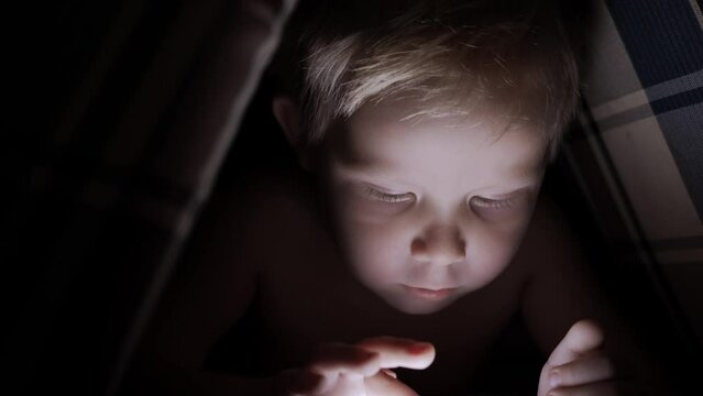 A little boy draws or watches cartoons on an electronic tablet in a pillow hut in the dark. The concept of children's interesting games and secrets. Close-up of a little boy's face. High quality 4k
