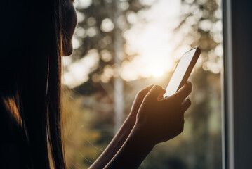 Close up profile side view of relaxed young woman sitting on comfy window uses smartphone on background autumn sunset and colorful forest, typing message, chatting with friends on social media