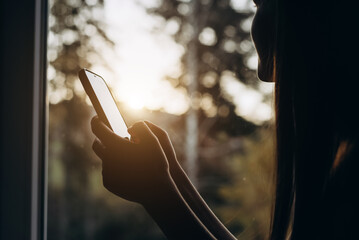Close-up of young caucasian woman holding smartphone sitting sitting on cozy window sill and enjoys scenic view on beautiful autumn forest during sunset or sunrise. Recreation and escaping to nature