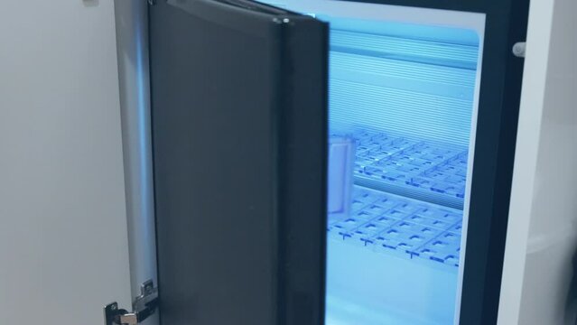 Door of empty minibar in hotel is slowly closing. World Food Crisis. Black trendy modern refrigerator in cozy room. Blue light inside fridge. Idle small icebox after cleaning.
