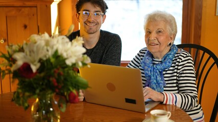Portraits of elderly senior woman and teen grandson working with laptop computer in a dining room.