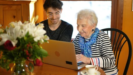 Teen grandson teaching elderly senior grandmother how to use a laptop computer in a dining room.