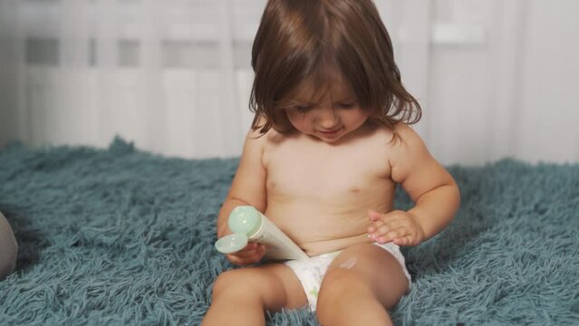 A cute two-year-old girl in a diaper applies cream to her legs