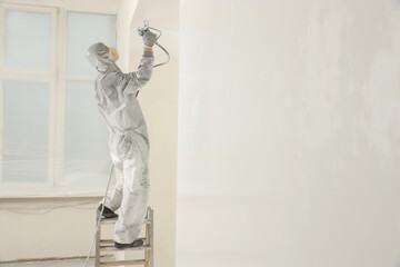 Decorator in protective overalls painting wall with spray gun indoors