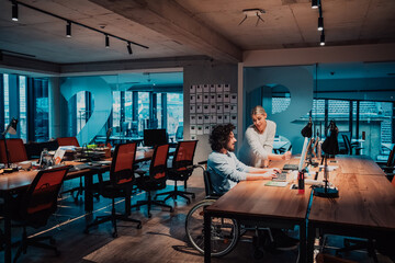 Fototapeta na wymiar A businessman in a wheelchair in a modern coworking office space working with a female colleague late at nigh. Colleagues in the background