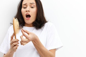 a very upset, scared, sad woman with long black hair stands in a white T-shirt on a light background and cleans a comb from fallen hair, expressing negative emotions on her face