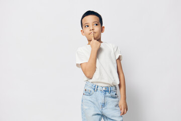  thoughtful boy, is standing in a white T-shirt on a light background with an empty space under the insertion of an advertising layout with a very thoughtful face, putting his finger to his mouth