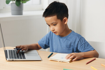 little boy, school age sits at a table with a laptop and makes notes in a notebook. Home schooling