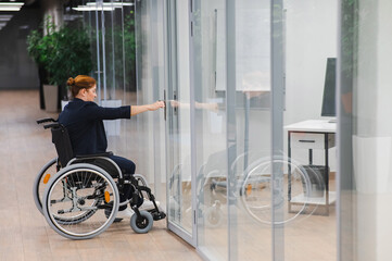 Red-haired caucasian woman in a wheelchair trying to open the door in the office.  - obrazy, fototapety, plakaty