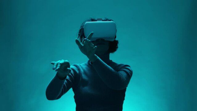 Mature Woman 40s Using Virtual Reality Headset In Studio on blue background. Adult Model Making Gestures to Reviewing Business Documents or Answering to Email Colleague.Modern Female on Online Meeting