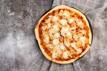 Homemade pizza with chicken and pineapple on a round wooden cutting board on a dark grey background. Top view, flat lay