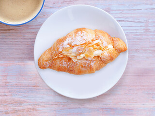 Top view, Croissant with cream and almonds on white plate and coffee cup over wooden table. Close up, space for text...