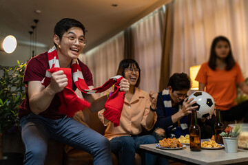 Group of Asian people friends sit on sofa watching and cheering football or soccer games competition on TV together at home.Happy man and woman sport fans celebrating sport team victory sports match