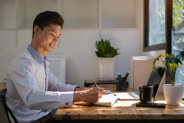 Start up businessman writing new idea on notepad and using laptop in bright modern office.