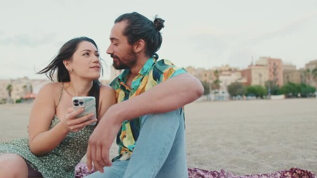Romantic couple uses mobile phone while sitting on the beach on buildings background