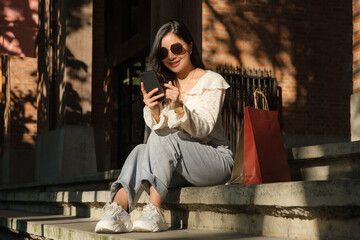 Beautiful woman using mobile phone, sitting on staircase after finished shopping in city square...