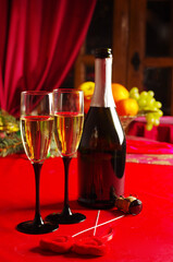 Two glasses of champagne, a bottle of champagne, a burning candle on a table with a red tablecloth.