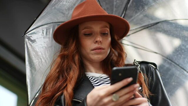 Close-up face of displeased redhead young woman in fashion hat using smartphone to order taxi on app standing with transparent umbrella in city street rainy weather outdoors. Shooting in slow motion.