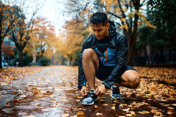 Young athlete tying shoelaces during rainy day in park. - obrazy, fototapety, plakaty