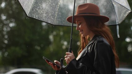 Side view of pretty young woman wearing fashion hat standing with transparent umbrella and dialing number using smartphone in city street. Elegant female talking on phone rainy weather outdoors. - Powered by Adobe