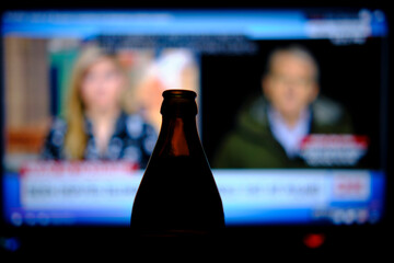 Drinking beer while watching the news. A bottle of beer is standing in the dark, in the background...