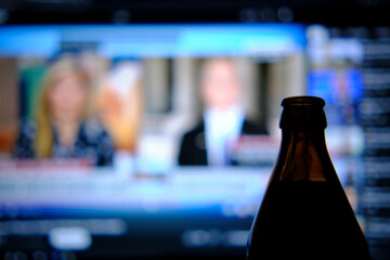 Drinking beer while watching the news. A bottle of beer is standing in the dark, in the background is a bright screen with the evening news