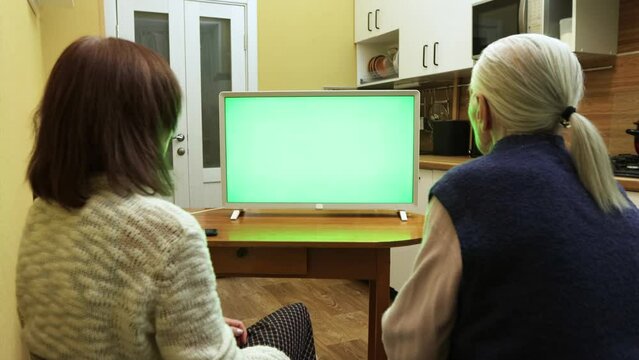 Family watching TV. Green screen. An adult daughter and grandmother are sitting at home in the kitchen and watching TV. In front of them is a green screen TV. 