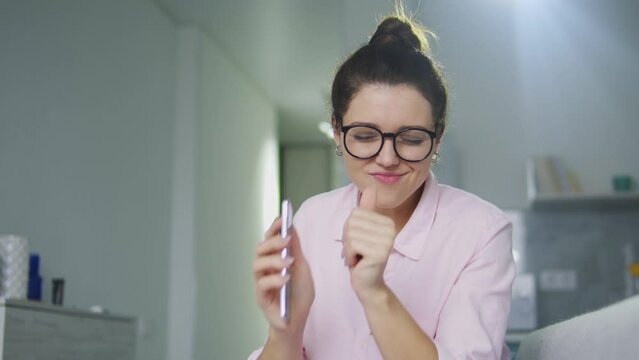 Cheerful excited woman in eyeglasses and shirt having fun, singing and dancing to music she turning on on her smartphone, free access to a large music library, subscription to music channels