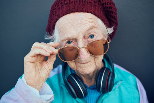 Senior Woman, Face And Fashion Sunglasses, Vintage And 90s Clothes, Cool Style And Headphones With Attitude Against A Wall Outdoor. Elderly Model, Edgy Attitude And Retro Portrait Or Retirement