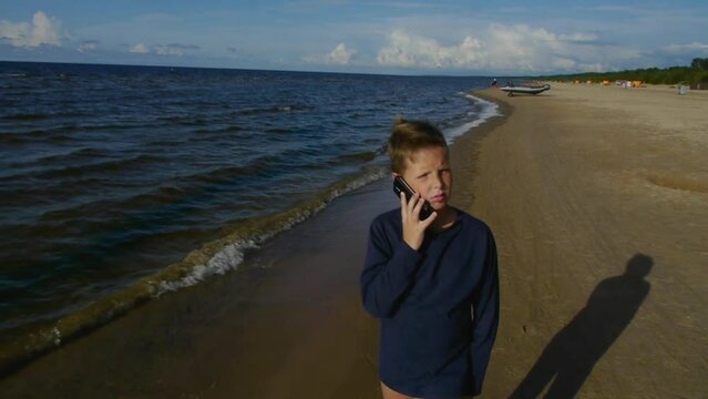 A teenage boy walks with his phone on the seashore.