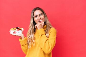 Young pretty Uruguayan woman holding a bowl of fruit isolated on red background happy and smiling