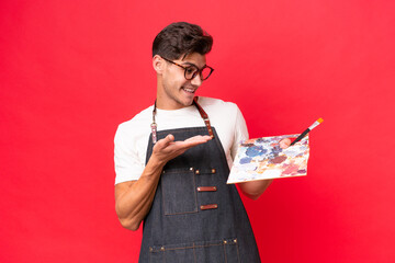 Young artist caucasian man holding a palette isolated on red background extending hands to the side...