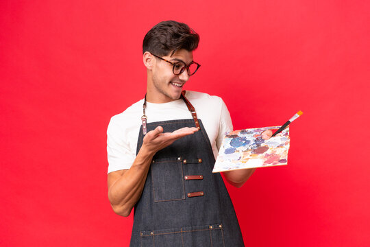 Young artist caucasian man holding a palette isolated on red background extending hands to the side for inviting to come
